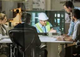 four business people sat at a desk watching a video of a worker on a television